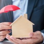 Businessman hand holding red Umbrella cover wooden Home model. real estate, insurance and property concepts